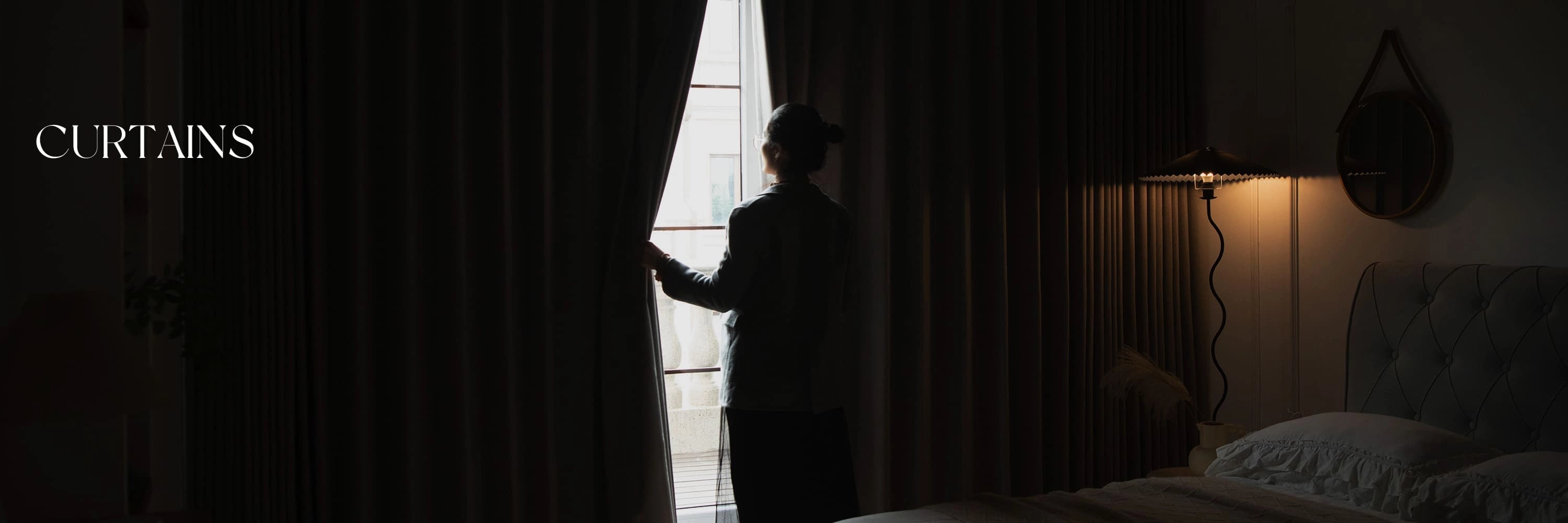 A woman is opening curtains in a dimly lit room with a bed and a lamp.