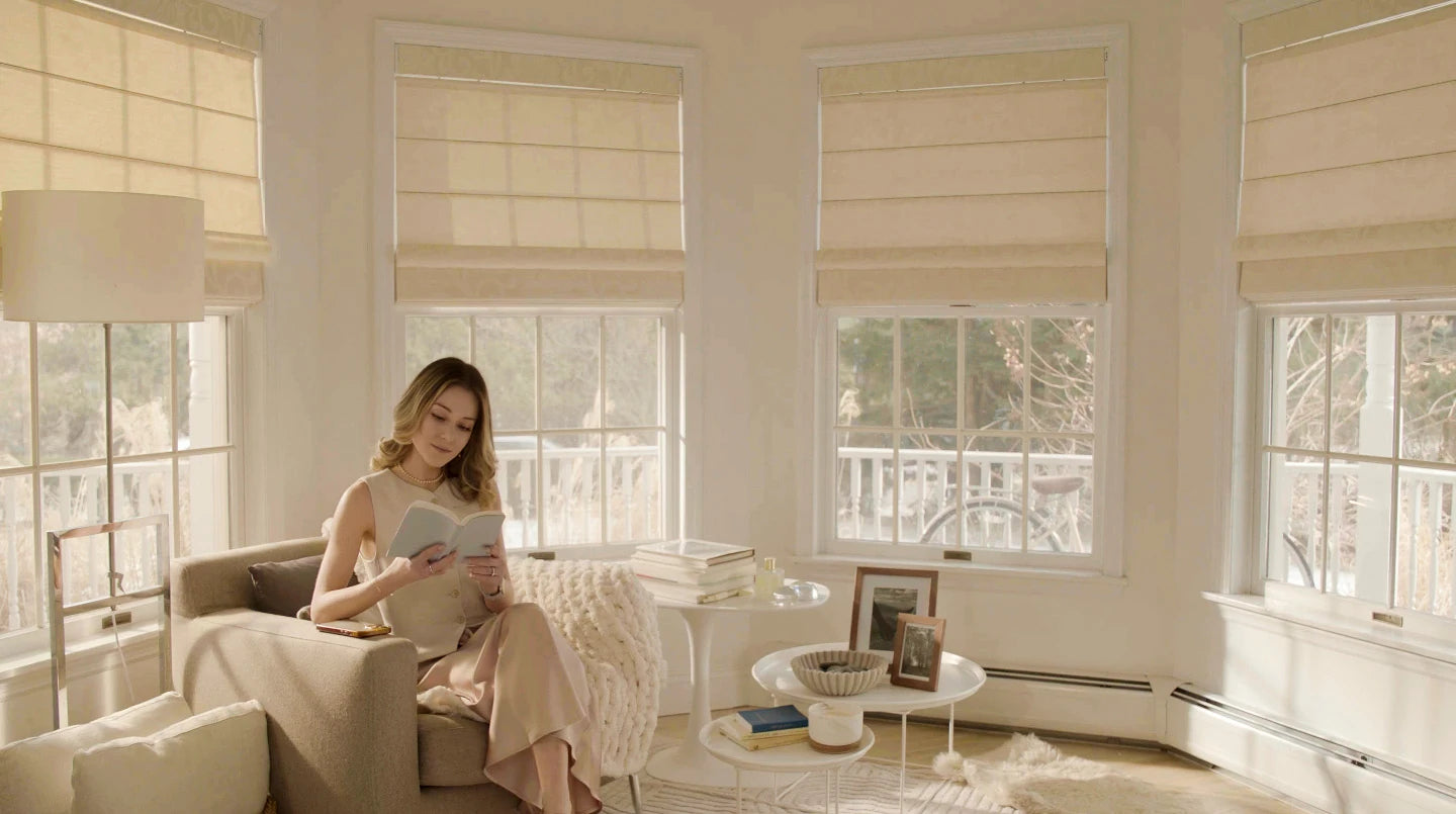 Woman reading a book in a bright room with large windows and beige blinds.