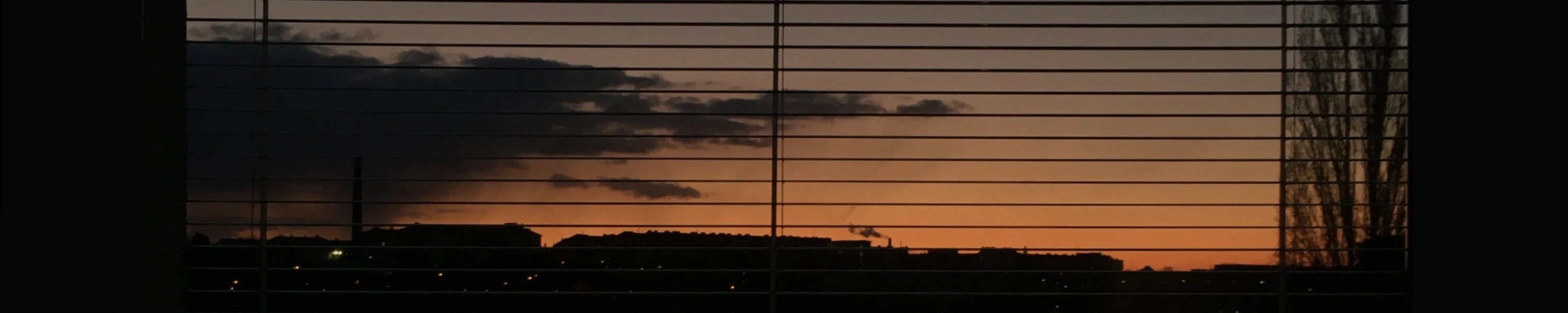 Sunset or sunrise through window blinds with silhouettes of buildings and trees.