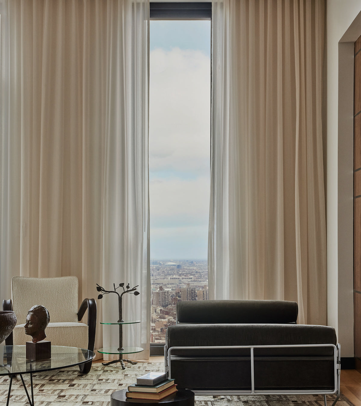 Modern living room with a large window covered by a curtain, offering a cityscape view, featuring a black sofa and a glass coffee table.