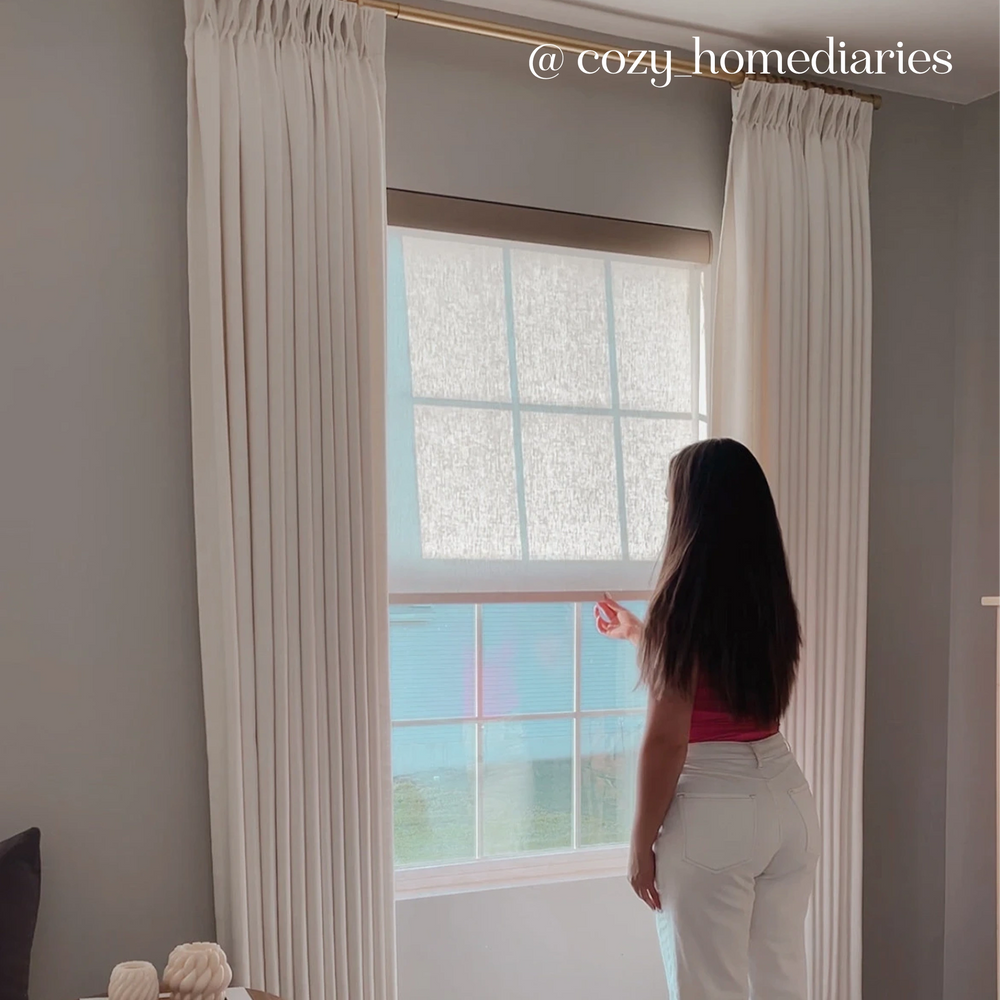 Person standing by a window with white curtains, looking outside.