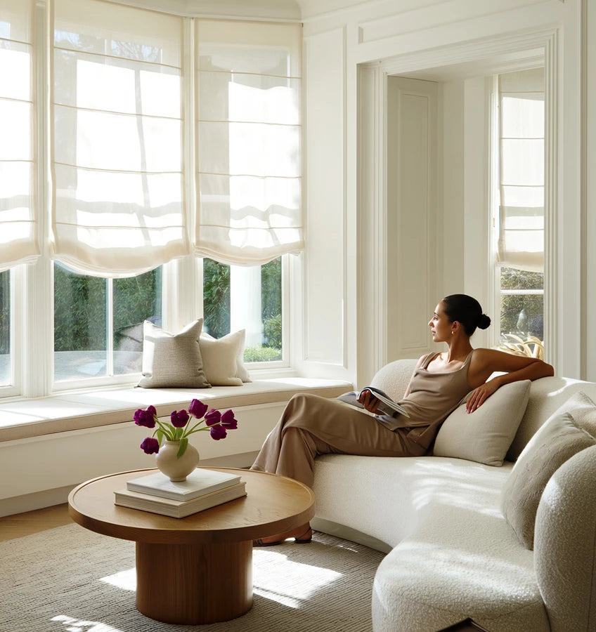 A woman sits on a white sofa in a bright living room with large windows and window treatments.