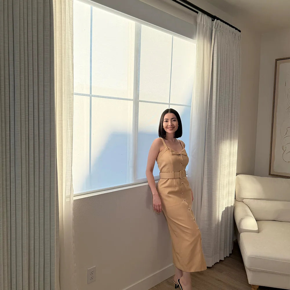 Woman in a beige dress standing by a window with dual shades in a room with a white sofa.