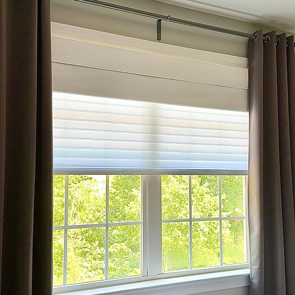Window with dual shangrila shades and brown curtains, showing greenery outside.