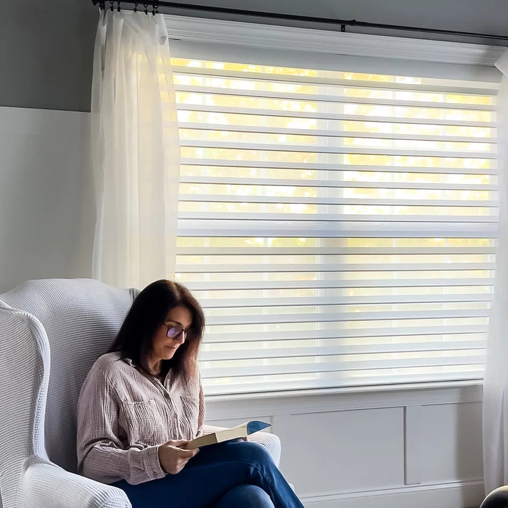 Woman reading a book in a chair by a window with white dual shangrila shades and curtains