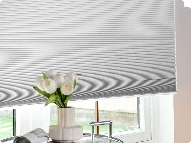 Window with cellular blinds and a vase of white flowers on a windowsill.