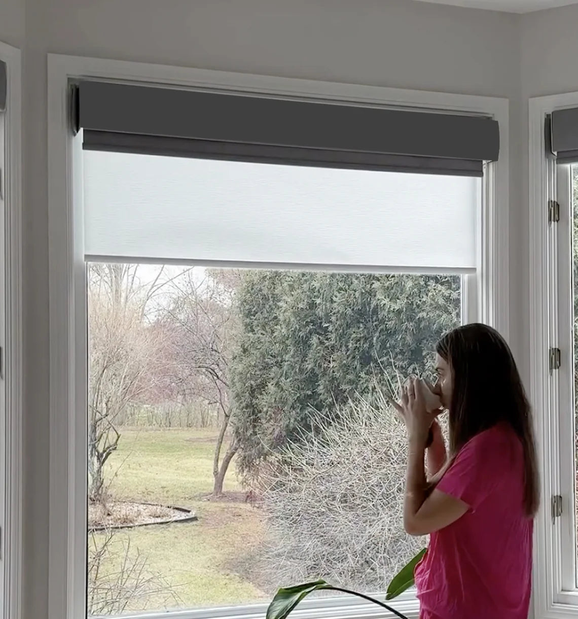 Person standing by dual roller shades with a view of trees and a garden, holding a cup.