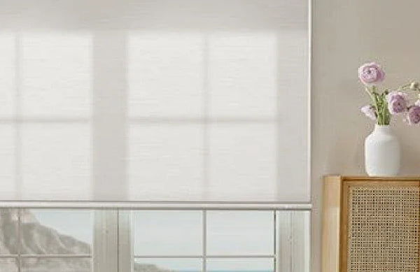 White roller blinds covering a window with a vase of flowers on a table to the right.