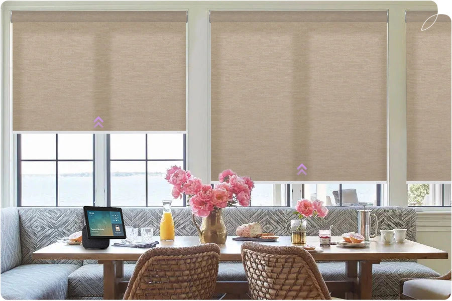 Beige roller blinds covering a window in a dining area with a table and chairs.
