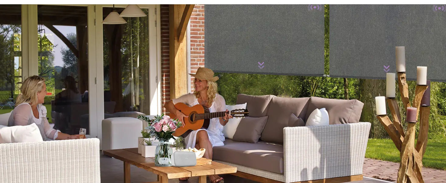 Woman playing guitar on a patio with a man and woman sitting on a sofa, surrounded by Graywind Outdoor Shades and decor.