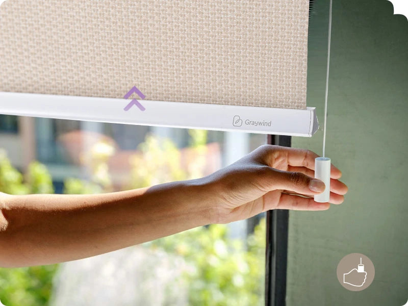Hand adjusting a roller blind with Graywind logo against a blurred outdoor background
