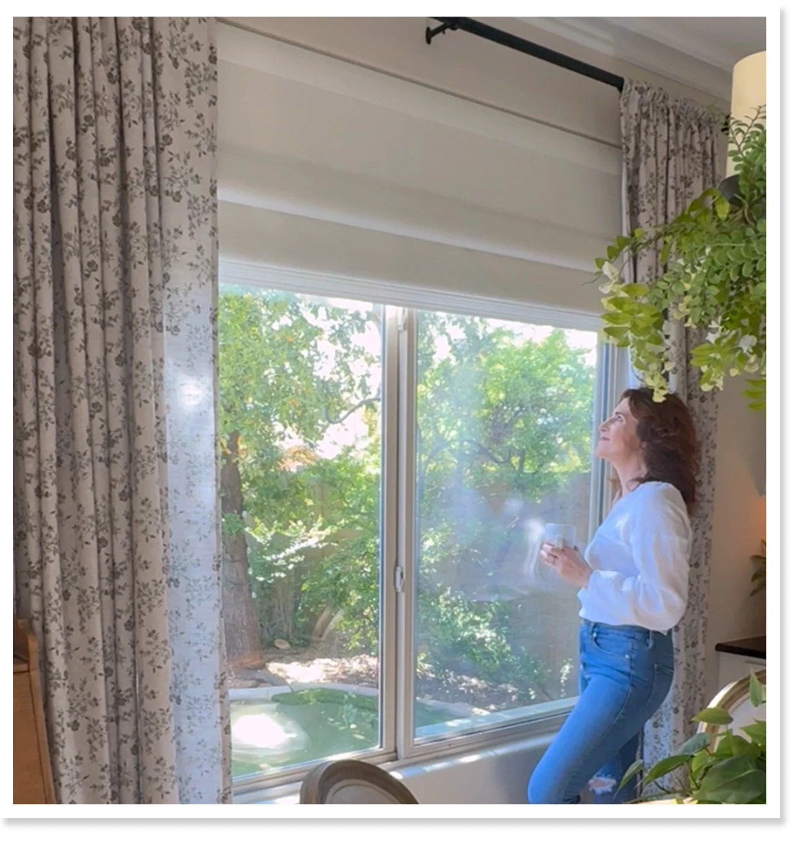 Woman standing by a window with roman shades and a view of trees and a pool.