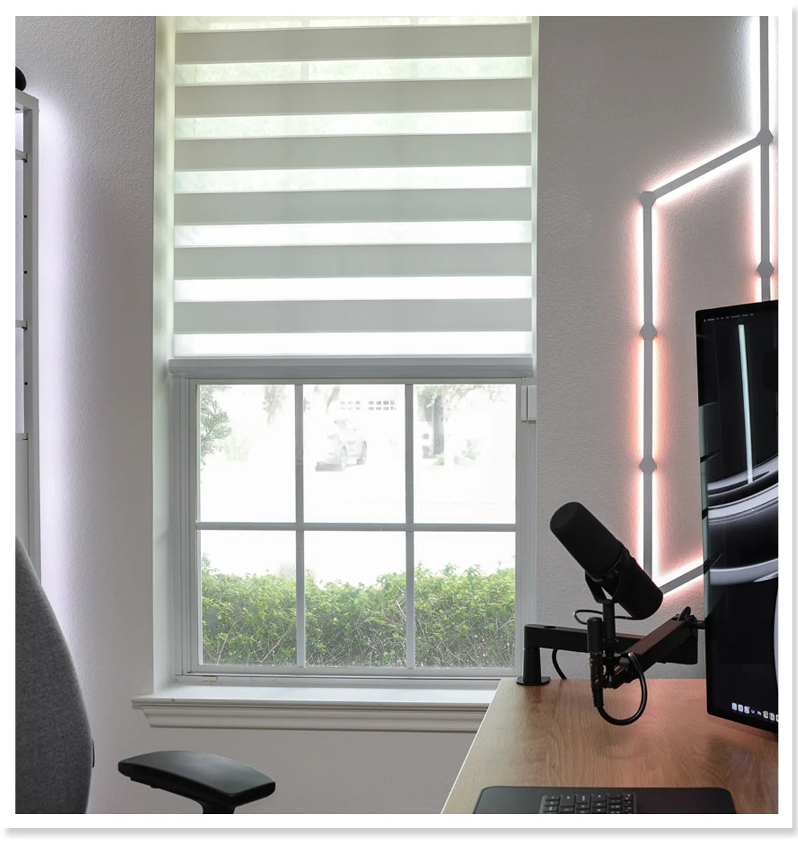 Home office setup with a desk, computer monitor, microphone, and chair near a window with zebra blinds.